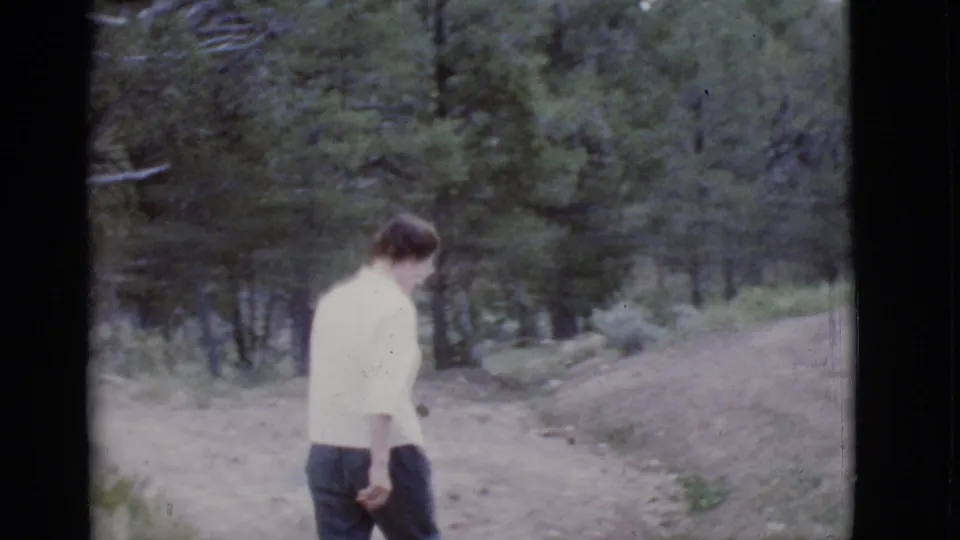 1968 Arizona 8mm frame showing a man walking along a dirt path through pine trees toward a scenic area.