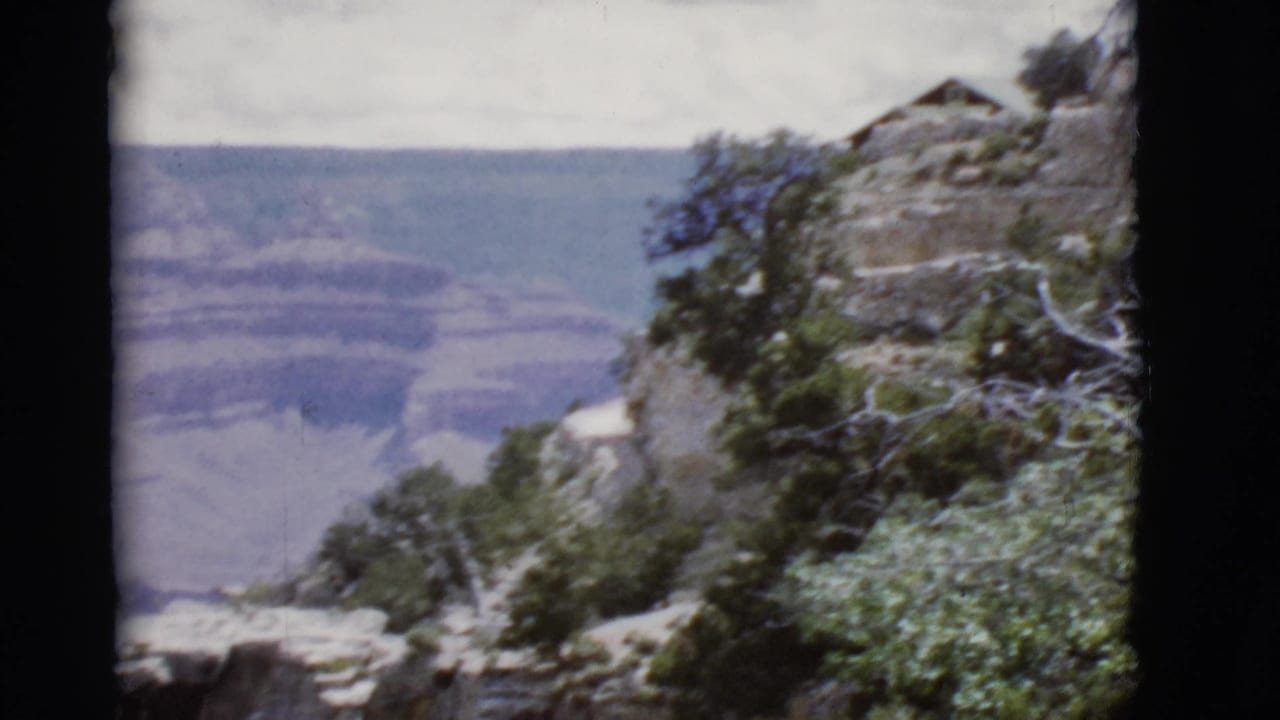 1968 Arizona home-movie frame showing a Grand Canyon rim cliff with shrubs, pale rocks, and a small structure above distant canyon layers.
