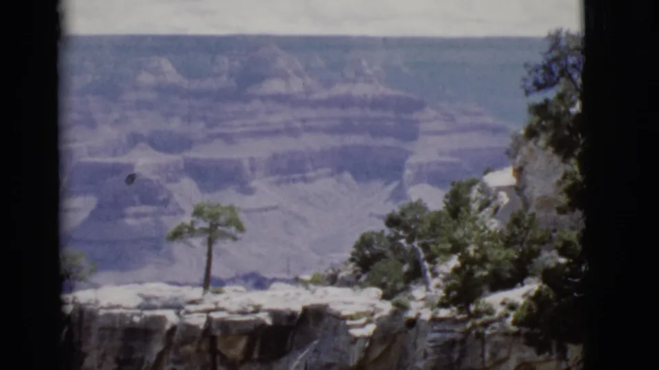 1968 Arizona 8mm frame of the Grand Canyon, with layered canyon walls behind a rocky rim, sparse shrubs, and a small tree.