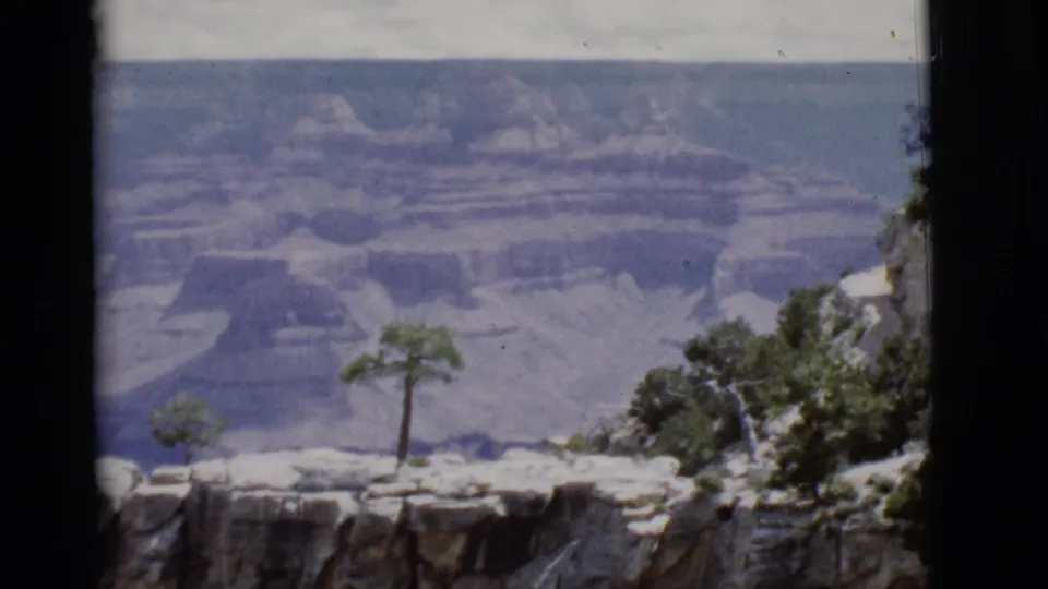 1968 Arizona 8mm frame showing a rocky Grand Canyon overlook, sparse trees, and hazy layered cliffs in the distance.