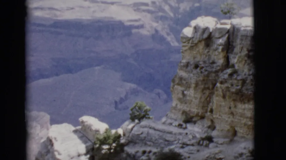 1968 Arizona home-movie frame showing a large cliff face above a blue-gray ravine with sparse trees on the rocks.