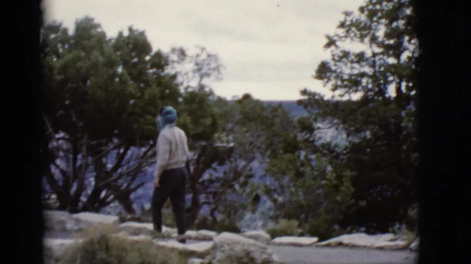 1968 Arizona frame of a woman in a blue scarf and light sweater walking beside green trees at the Grand Canyon overlook.