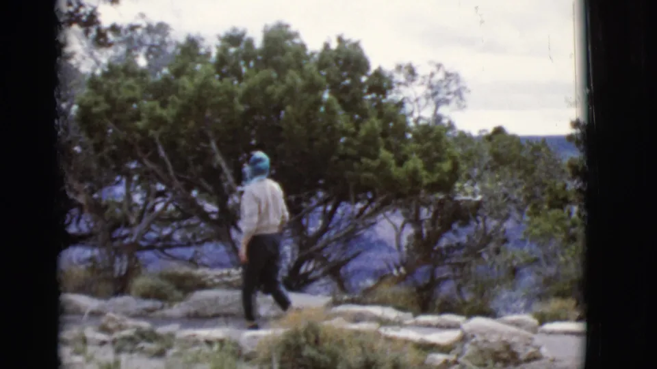 1968 Arizona 8mm frame showing a woman from behind on a stone path, with green trees and canyon walls beyond.