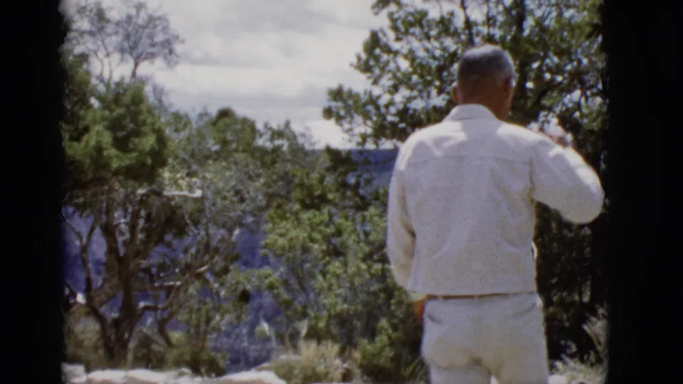 A man in a white shirt and light pants faces away toward an Arizona canyon view, framed by trees and 8mm film edges.