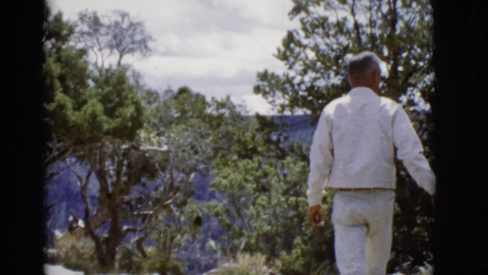 The man begins walking away across the overlook in 1968 Arizona footage, with trees and blue canyon forms in the distance.