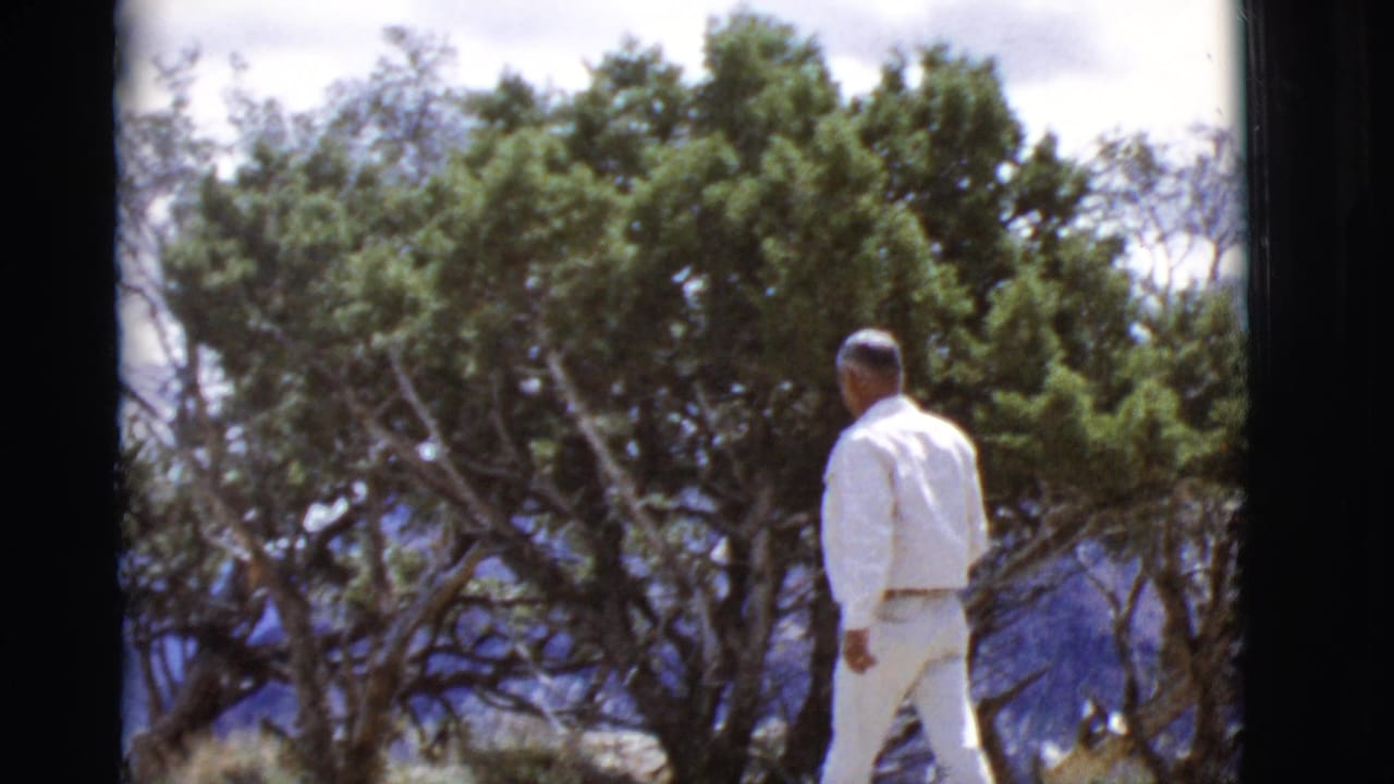 Man in white clothing walks past trees with mountains behind him.