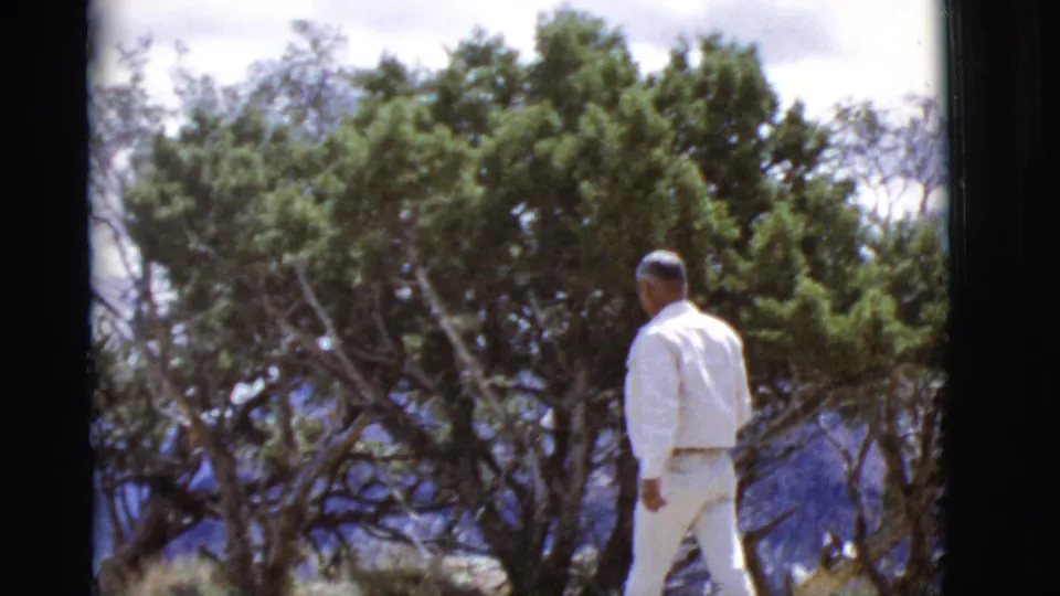 A man in white clothing walks along an Arizona overlook, with trees and distant mountains behind him.
