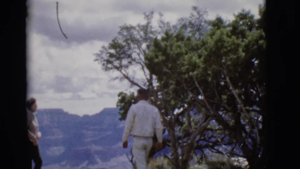 A man in white clothing walks away near trees, while another adult briefly appears at the edge of the Arizona overlook frame.