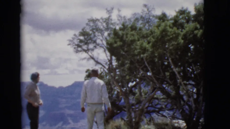 Two older adults in 1968 Arizona home-movie footage look across a canyon from a scrubby overlook beside a large tree.