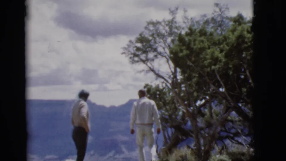 The older couple move along the Arizona canyon overlook, seen from behind in soft 8mm color with a tree at right.