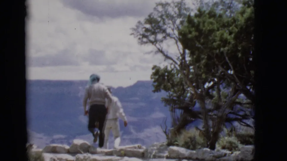 Two adults in 1968 Arizona walk near a rocky canyon overlook, with a tree and distant canyon walls beyond them.