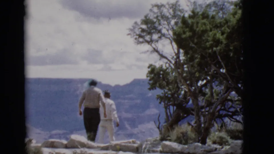 The couple continues along the Arizona canyon overlook, seen from behind with rocks, trees, and distant mesas in view.