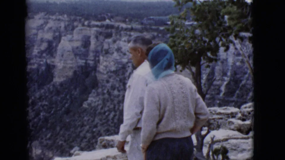 At the Arizona canyon overlook, the couple stands near the edge; the woman's blue headscarf and white sweater are clearly visible.