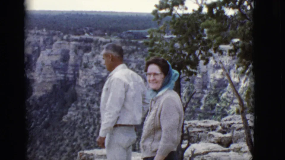 1968 Grand Canyon home-movie frame showing an adult couple at the rim, the woman smiling beside trees and layered rock walls.
