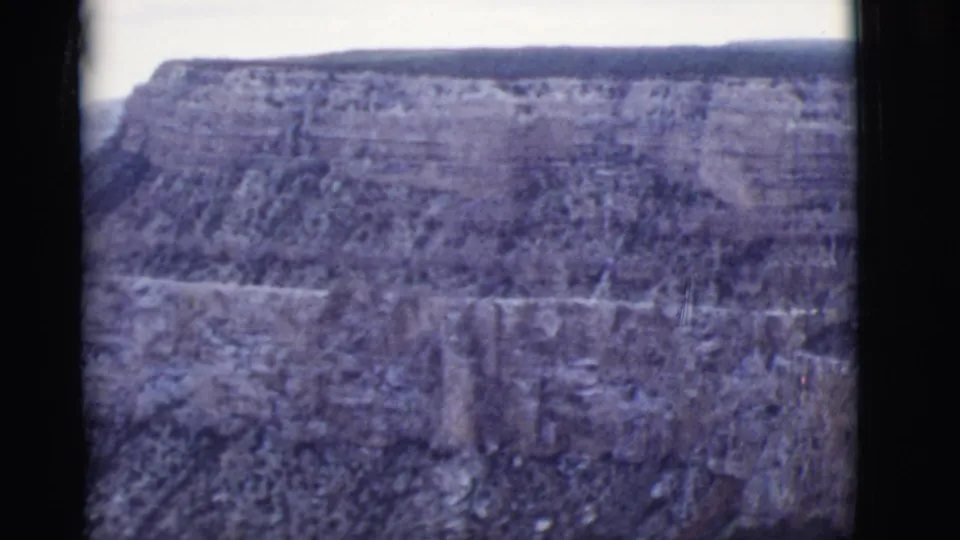 1968 Arizona 8mm frame showing a broad canyon wall, layered rock ledges, and a flat-topped mesa in soft blue-purple color.