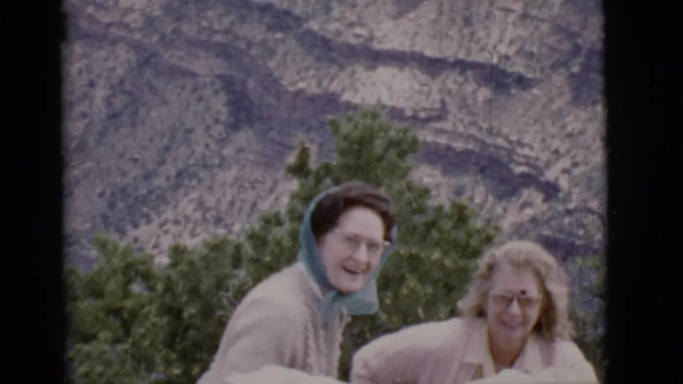 1968 Arizona home-movie still of two women smiling near green trees, with layered rocky terrain rising in the background.