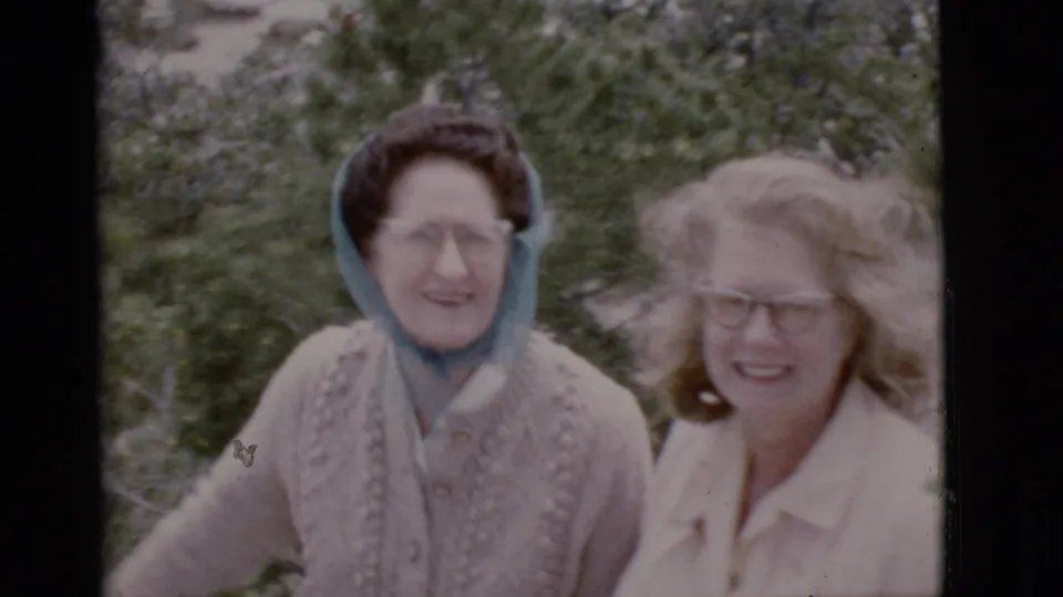 Two adult women in 1968 Arizona footage smile toward the camera, one wearing a blue headscarf and sweater, the other in glasses and a light blouse.