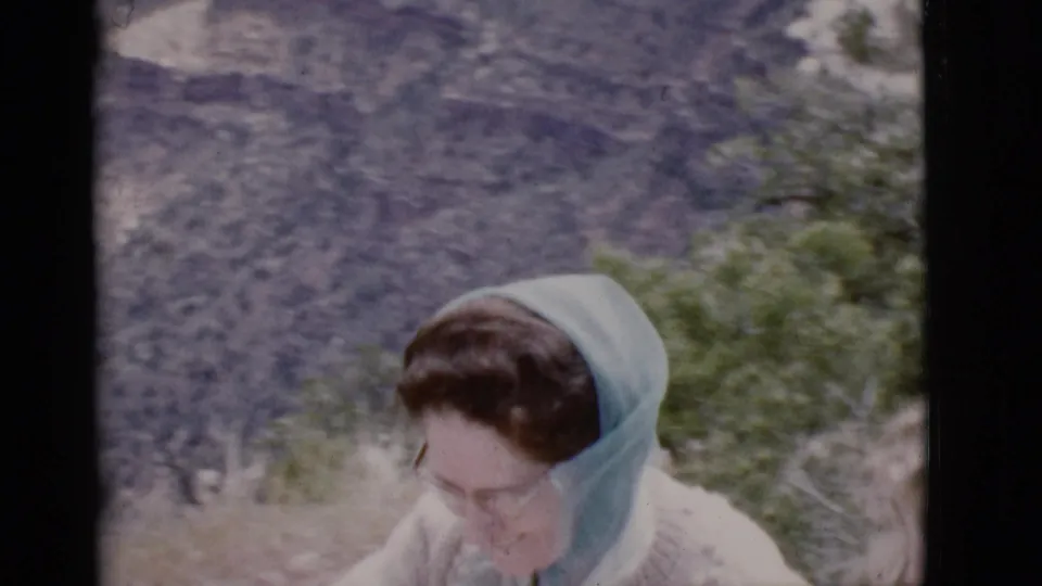 A woman in a pale blue headscarf and light sweater appears in soft 8mm focus against an Arizona canyon backdrop.