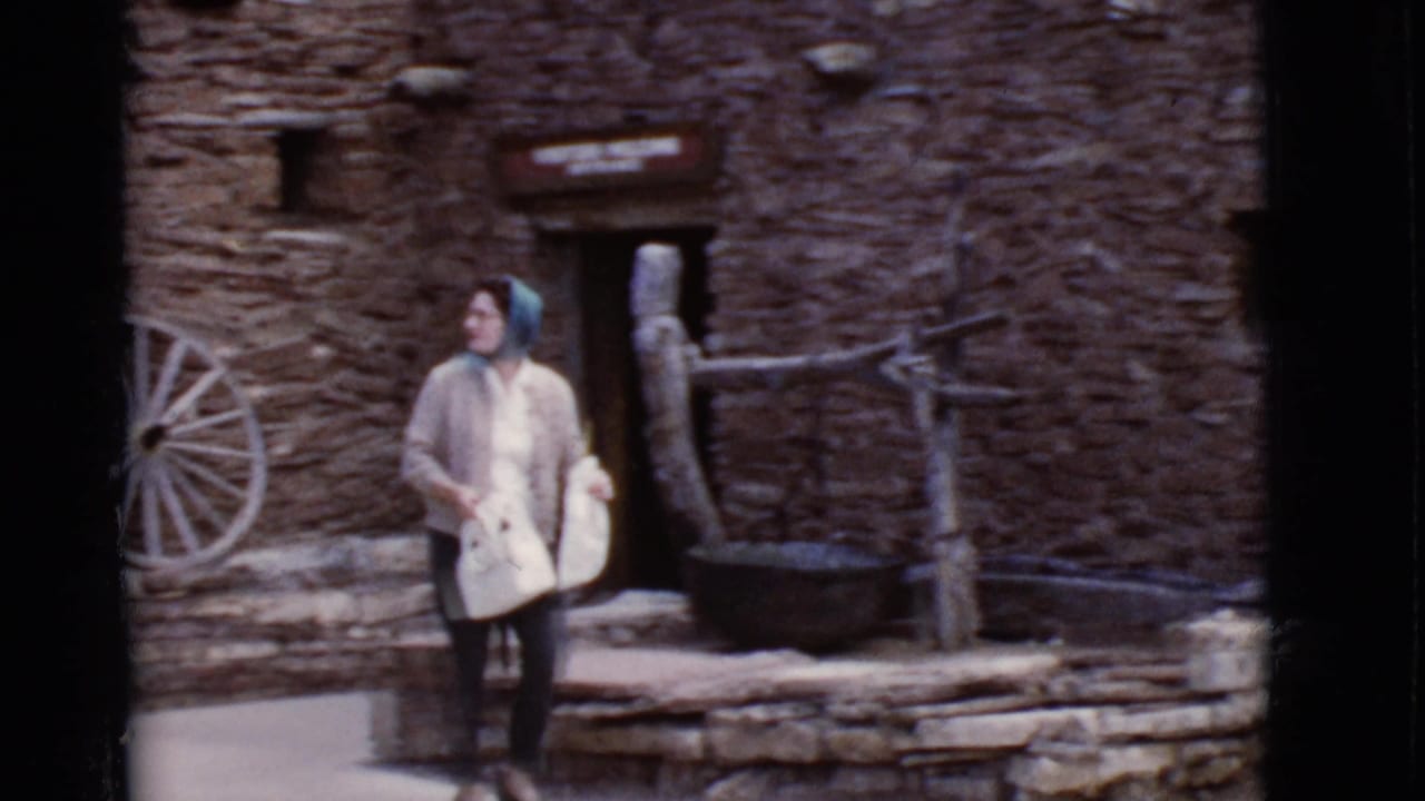 Woman in a cardigan and headscarf stands outside a stone building near a wagon wheel.