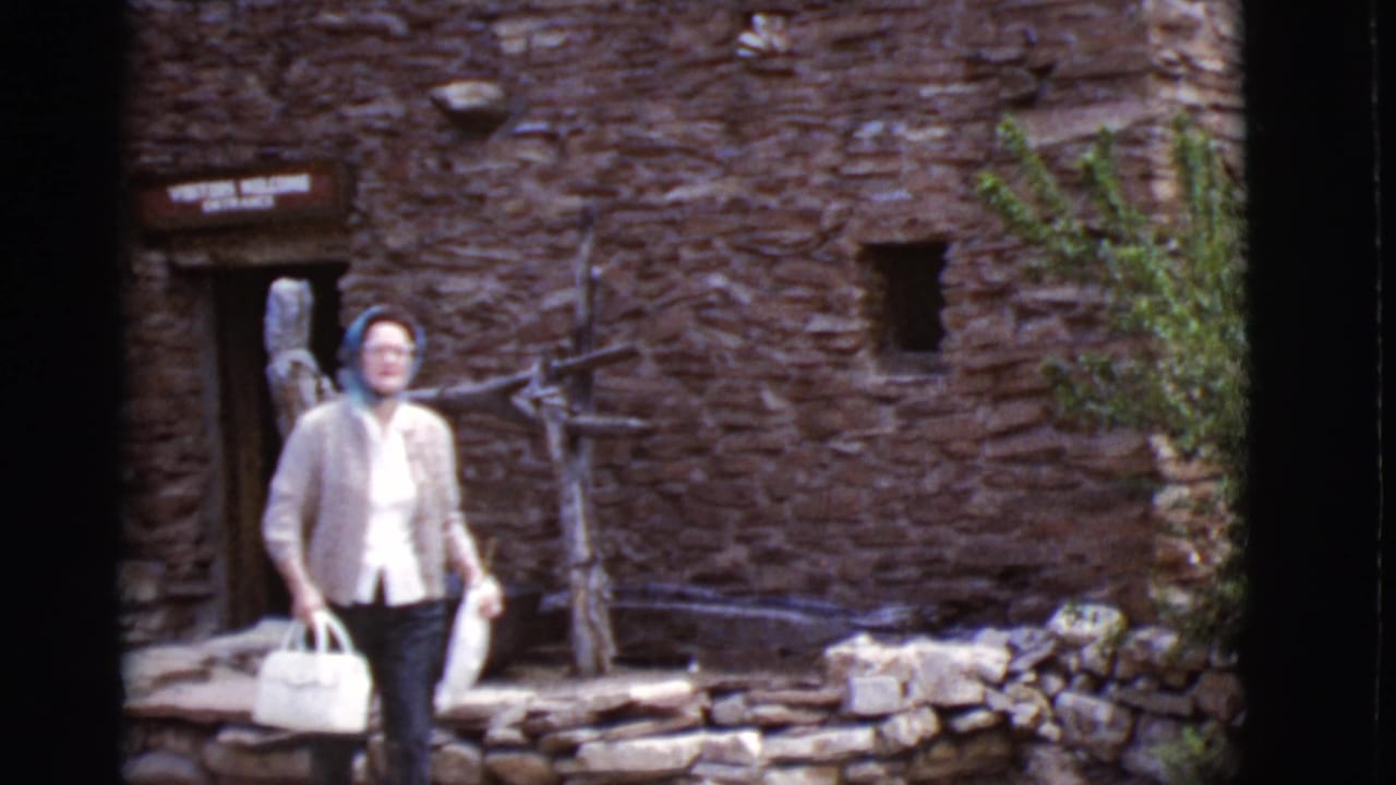 The woman walks forward outside the Arizona stone building, carrying a white handbag with rustic fencing and stonework behind her.