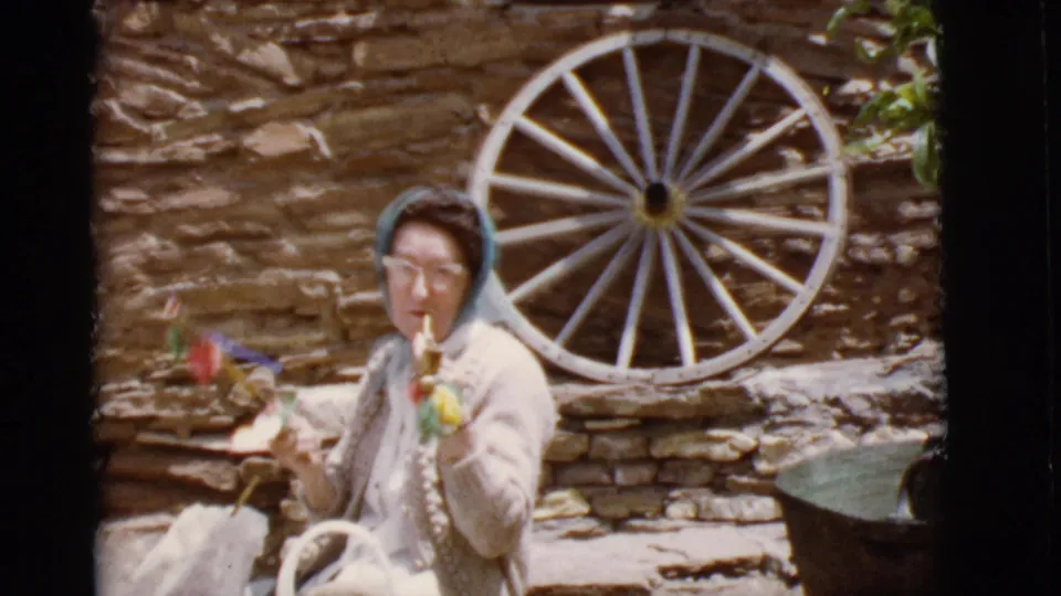 A closer 1968 Arizona frame shows the woman facing the camera near a large wooden wagon wheel and stone wall, with colorful items in her hand.