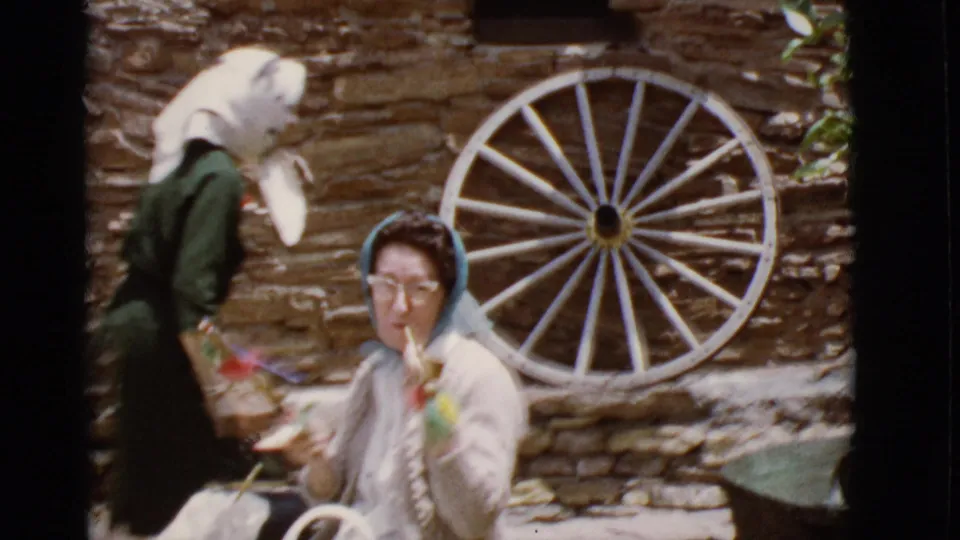 The woman remains near the wagon wheel and stone wall while another adult in a head covering passes through the foreground.