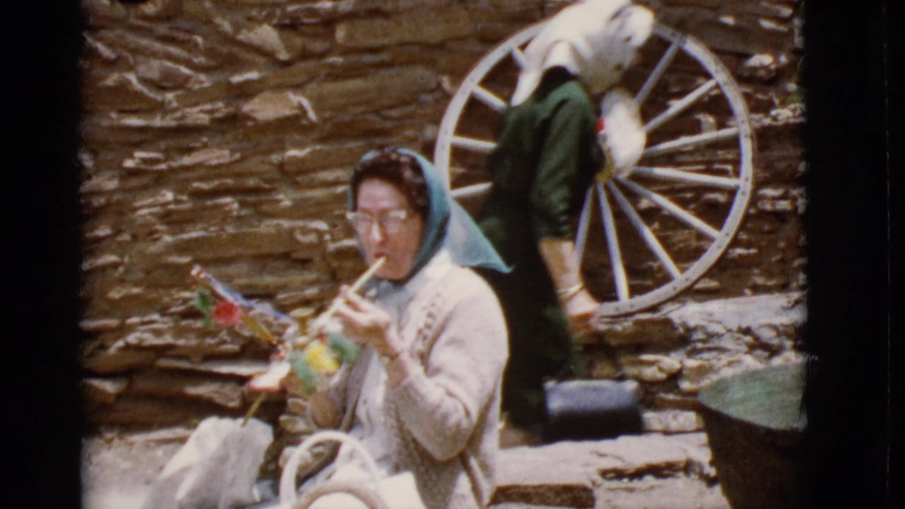 Elderly woman in a blue headscarf sits with colorful crafts beside a stone wall and wagon wheel.