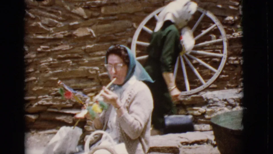 1968 Arizona home-movie frame of an elderly woman seated with handmade crafts near a rustic stone wall and white wagon wheel.