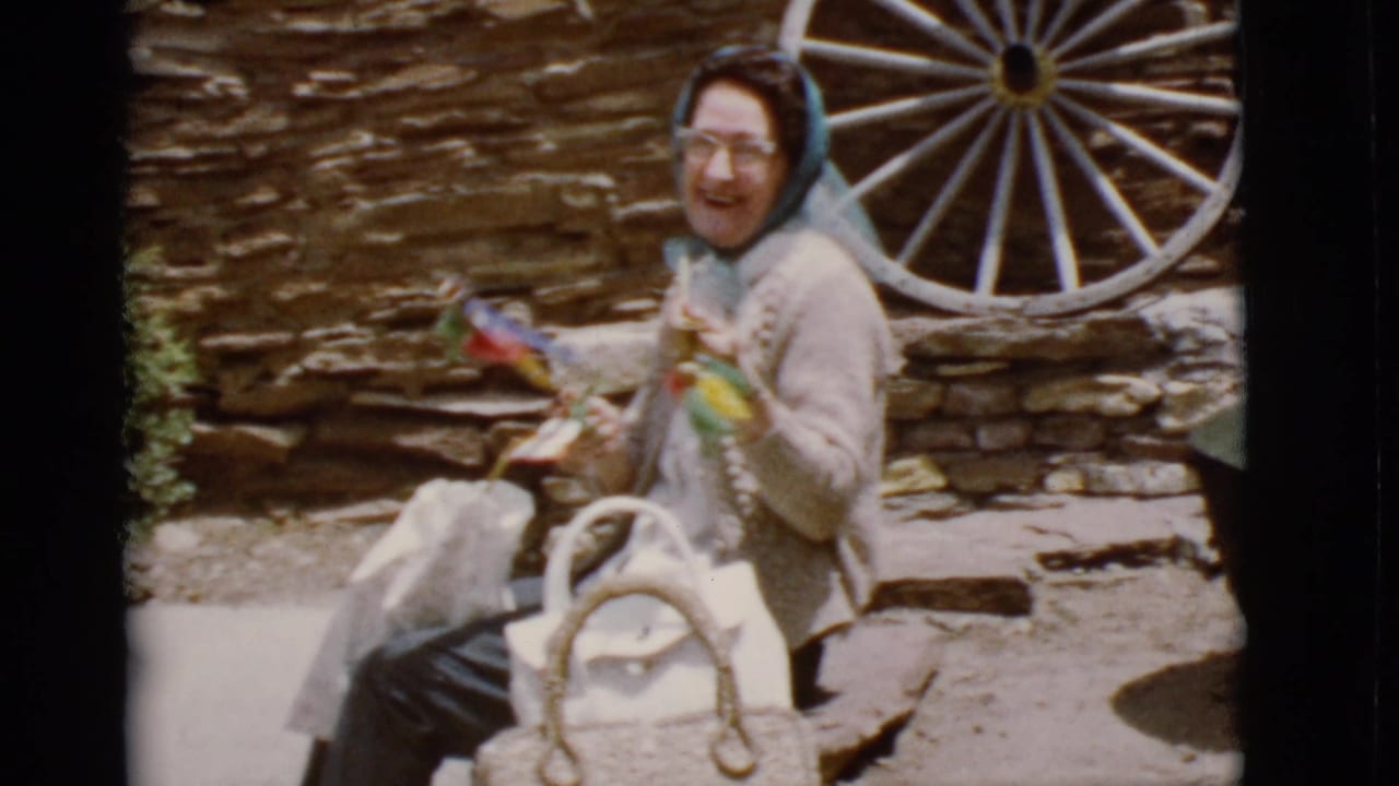 Smiling elderly woman holds colorful handmade items while seated near a white wagon wheel.