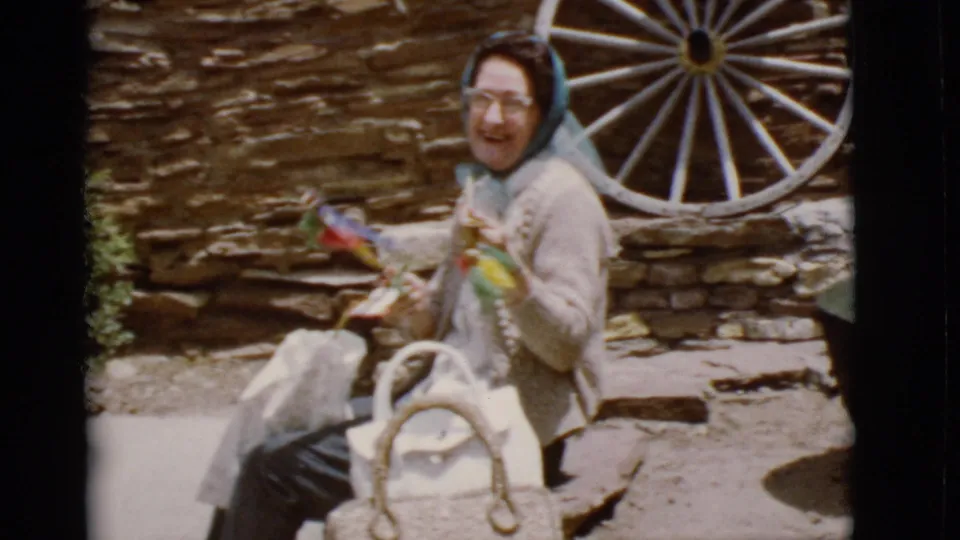 1968 Arizona home-movie frame showing a smiling elderly woman with handmade crafts beside a stone wall and wagon wheel.