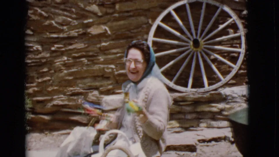 1968 Arizona home-movie frame of the seated woman turning toward the camera with colorful crafts near a wagon wheel.