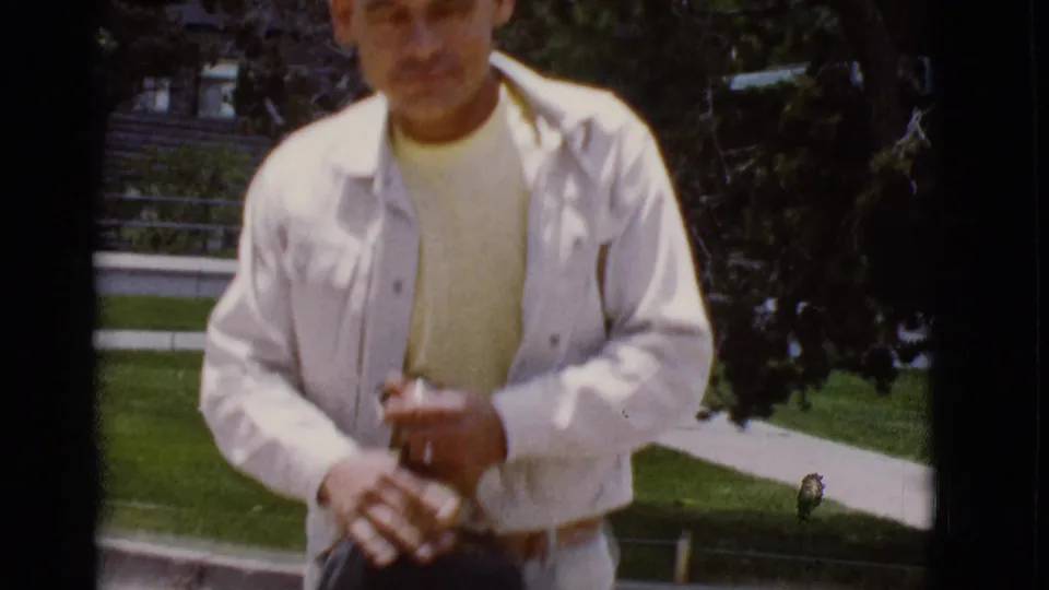 1968 Arizona home-movie frame of a man in a white jacket standing near grass and a sidewalk while adjusting a small bag.