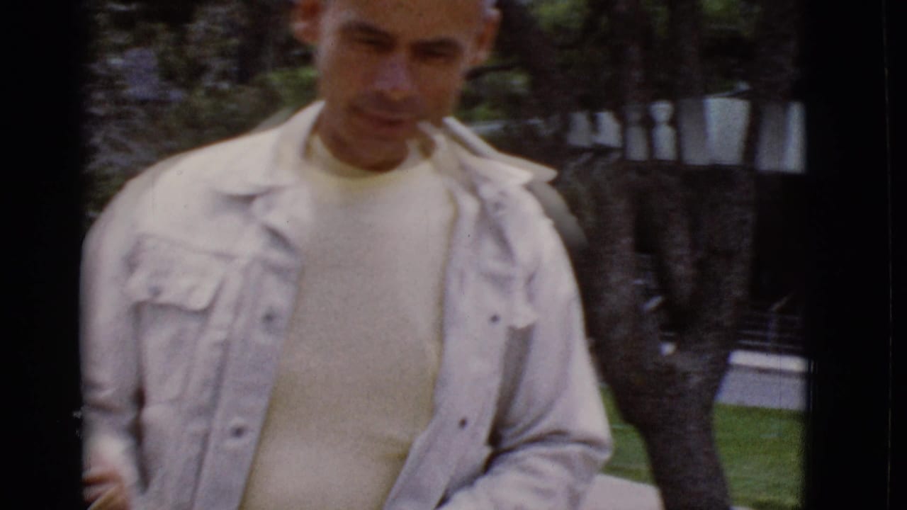 Man in a light jacket standing close to the camera near trees and grass.
