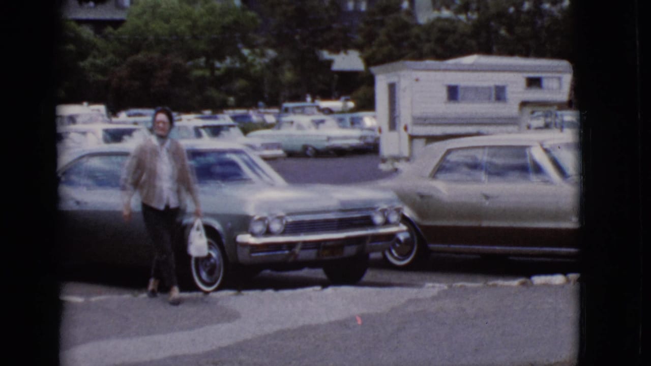 Woman carrying a white bag walks through a parking lot between parked cars.