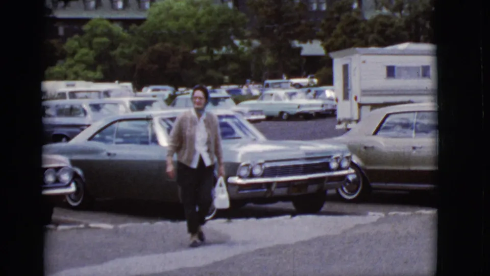In the supplied 1968 Arizona home-movie clip, a woman walks toward the camera past large parked cars and a camper trailer.