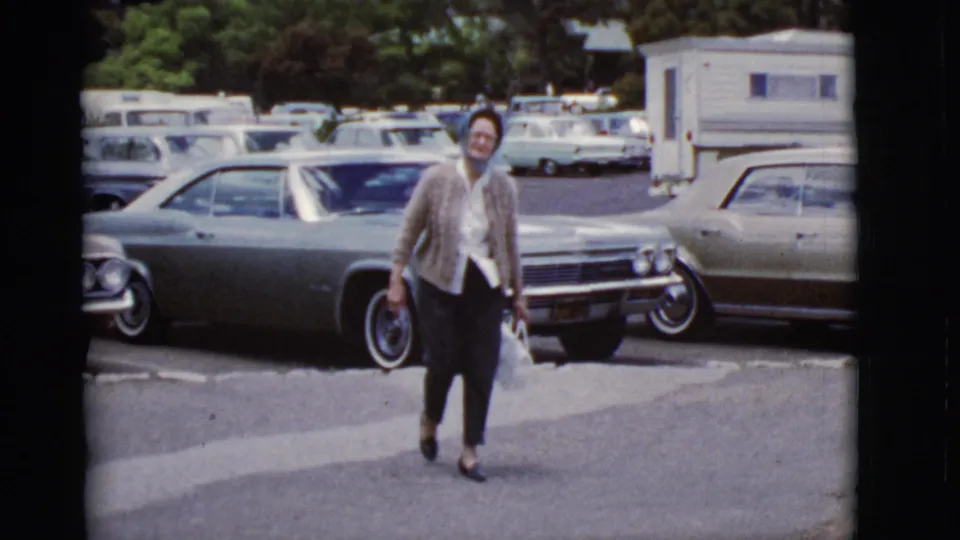 Woman in a cardigan, light blouse, and dark pants nears the camera in a parking lot with 1960s-era cars visible behind her.