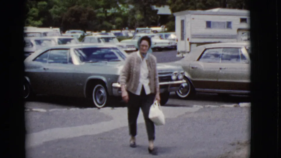 In 1968 Arizona home-movie footage, a woman in a headscarf and cardigan walks toward the camera in a lot with parked classic cars.