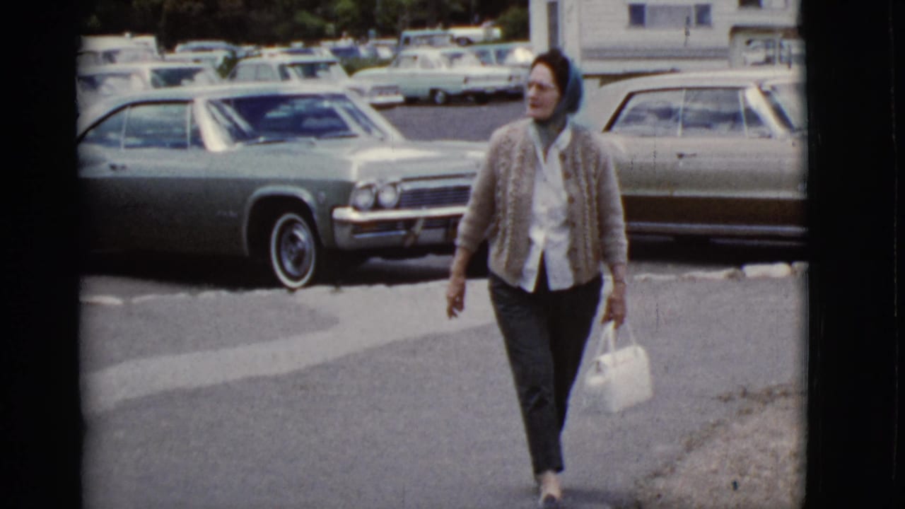 Woman carrying a handbag walks past parked cars in a paved lot.