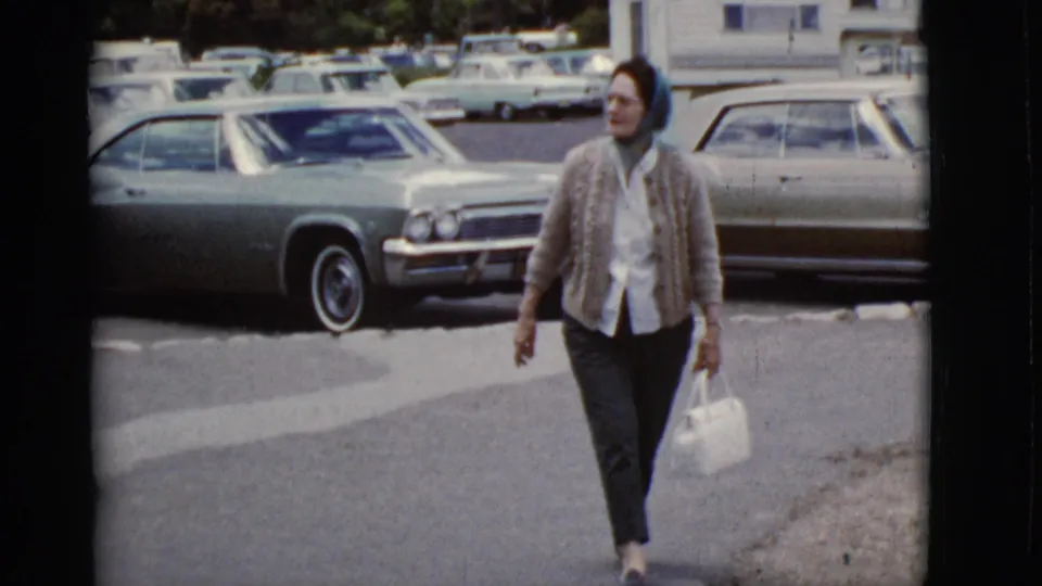 A woman in a blue headscarf and light cardigan carries a white handbag through a 1968 Arizona parking lot with large period cars.