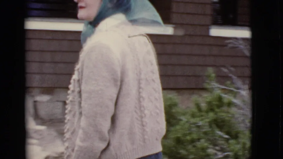 A close side view shows the woman’s headscarf, glasses, and cable-knit cardigan as she passes shrubs near a brick building in Arizona.