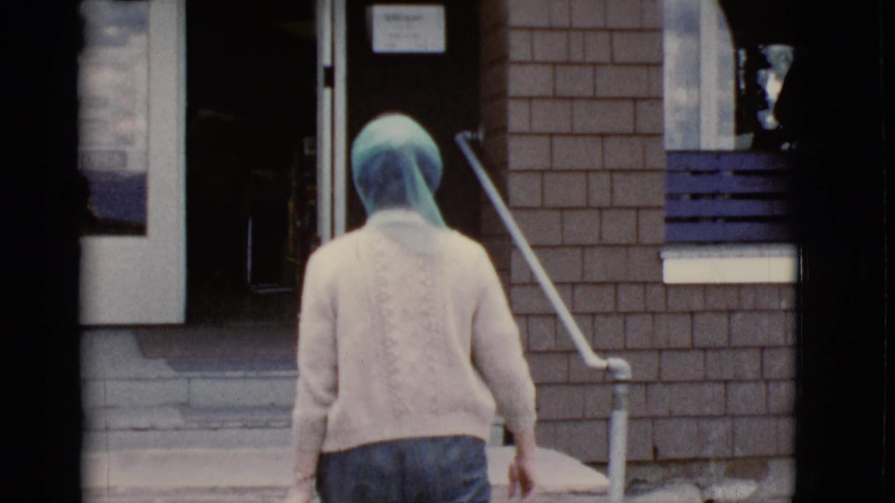 Woman in a blue headscarf walks up steps toward a glass door beside a brick wall.