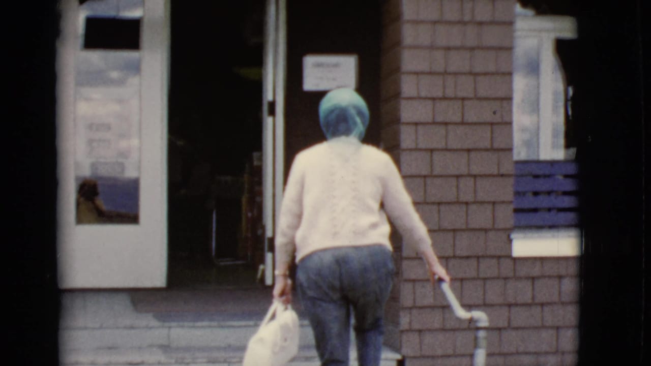 The woman reaches the entrance area with a white handbag, stair rail, glass door, and brick facade visible.