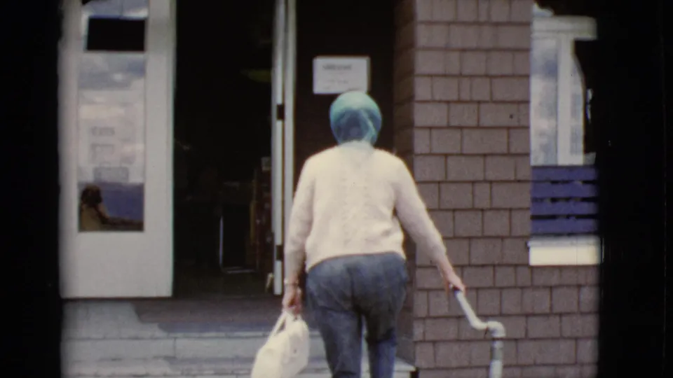 The woman reaches the entrance area with a white handbag, stair rail, glass door, and brick facade visible.