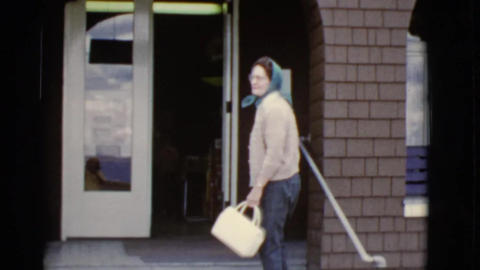The woman turns back toward the camera near the glass door, her blue headscarf and white handbag visible.