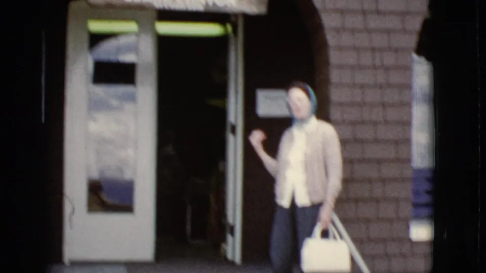At the doorway, the woman gestures back toward the camera before entering the brick tourist building.