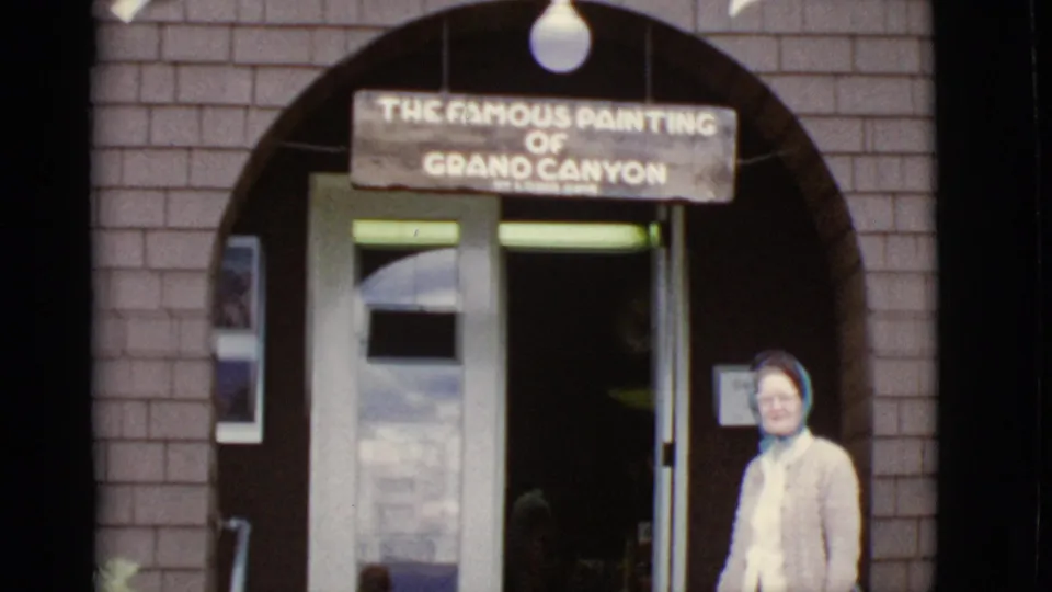 1968 Arizona home-movie frame showing a woman outside a brick building under a sign reading the famous painting of Grand Canyon.