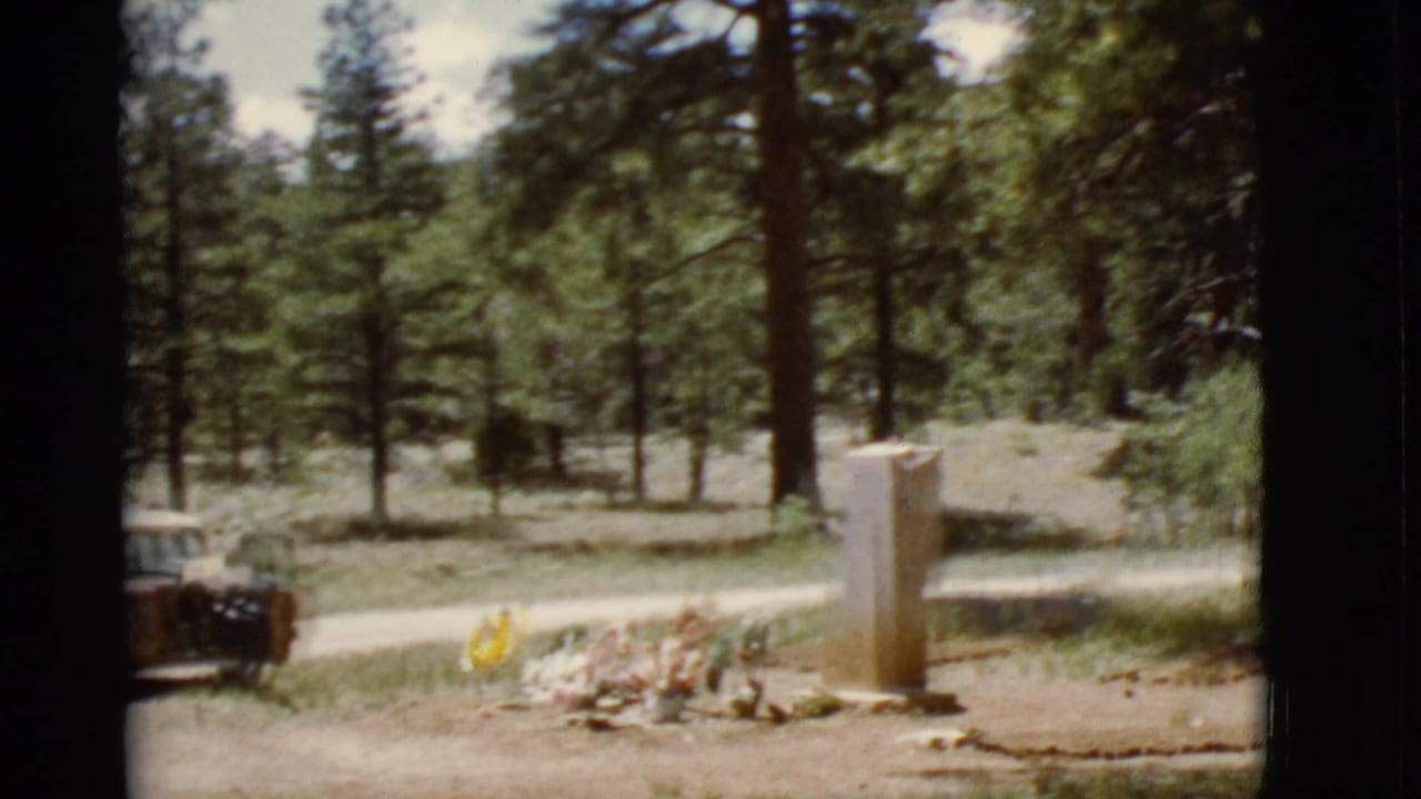 Forested clearing with a stone marker and floral tributes on the ground.