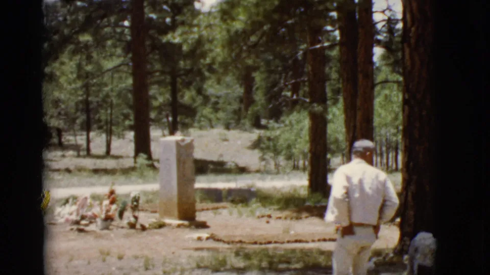 1968 Arizona home-movie frame showing a man in light clothing near a wooded marker with flowers at its base.