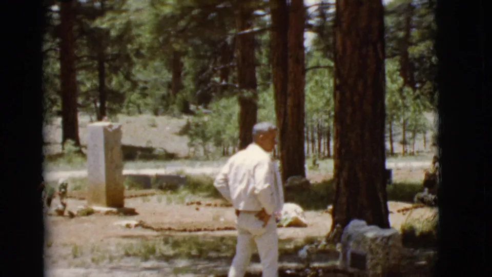A man in light clothing stands among pine trees near a stone marker and floral tributes in the 1968 Arizona footage.