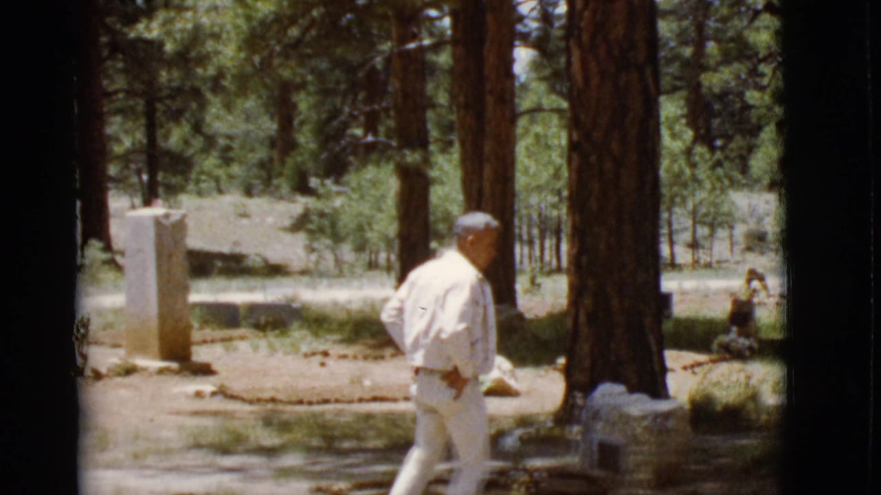 Man in a light suit walking past grave markers among tall pine trees.